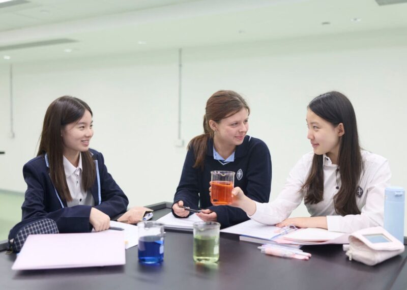 Three girls sat at a desk with a book and a beaker each.